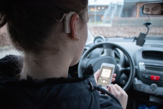 Bild mit Frau im Auto hält ein Smartphone. Cochlea-Implantat (CI) und Hörgerät mit nur einer App regeln –– Cochlear und ReSound ermöglichen bimodale Steuerung über die Nucleus® Smart App (Foto: Smart Hearing Alliance) 
